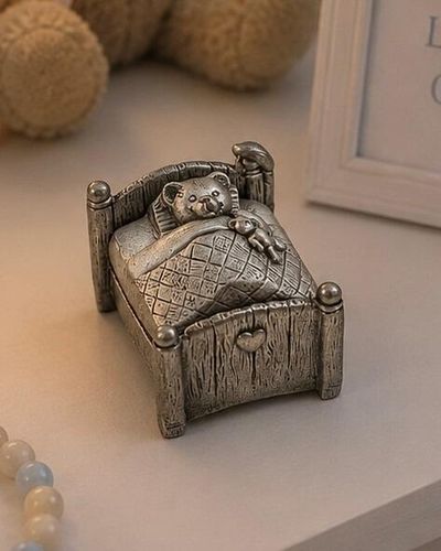 Pewter keepsake box sculpted in the shape of a miniature wooden bed, featuring a teddy bear tucked under a quilted blanket lid holding a tiny toy bear, displayed with nursery decor
