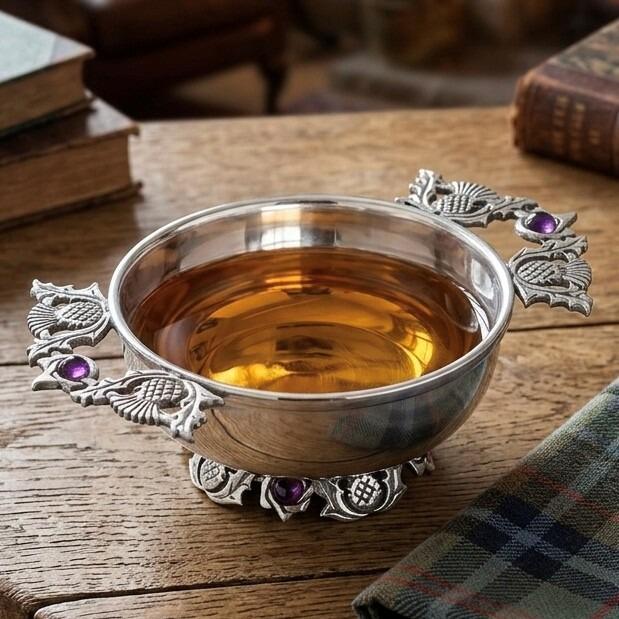 Traditional pewter quaich bowl filled with whiskey, featuring sculpted Scottish thistle handles and a decorative base inlaid with purple gemstones, displayed on a wooden table alongside a tartan cloth