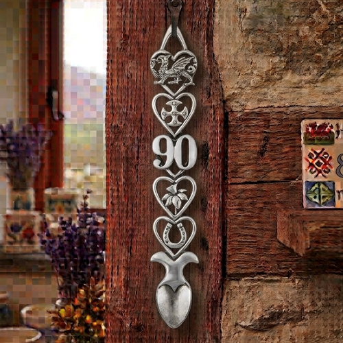 Handcrafted pewter 90th birthday Welsh lovespoon hanging on a rustic wooden beam, featuring detailed symbols including a Welsh dragon, Celtic cross, the number 90, a daffodil, a horseshoe, and a heart-shaped spoon bowl.