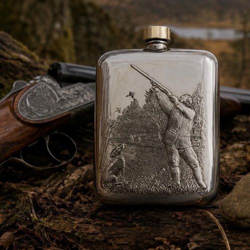 A polished pewter hip flask with a smooth brass screw-top cap, resting on a mossy, textured log outdoors. The front of the flask features an intricate, embossed relief scene of a hunter aiming a shotgun upward at flying birds, with a gundog sitting on the ground nearby amidst trees and grass. An engraved, break-action shotgun lies softly out of focus in the background