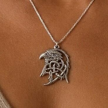 A close-up photograph of a pewter Celtic eagle head pendant suspended from a silver chain and worn against the skin. The detailed openwork design depicts an eagle profile formed by interwoven traditional Celtic knotwork, which also comprises the feathers, with a dark, antiqued finish