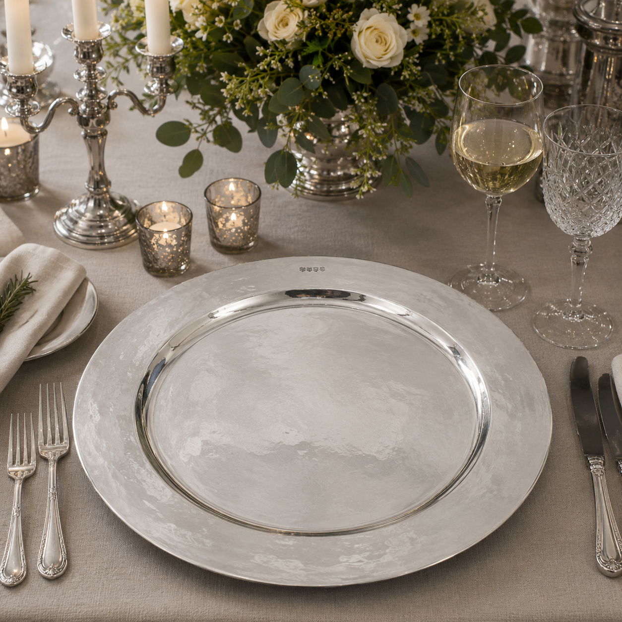 Handcrafted pewter 19 inch charger plate with polished hammered finish, luxury dining table centrepiece displayed with crystal glassware, silver cutlery, candlelight and elegant floral arrangement