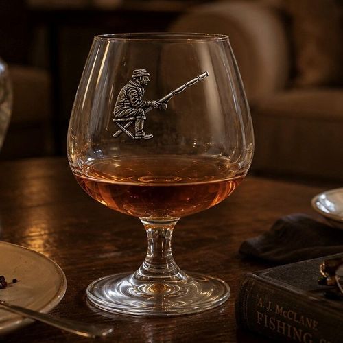 A short-stemmed clear glass brandy snifter filled with amber brandy, resting on a rustic wooden table. The front of the glass bowl features a detailed pewter relief emblem of a fisherman wearing a hat, seated on a folding stool and angling with a fishing rod. In the foreground to the right