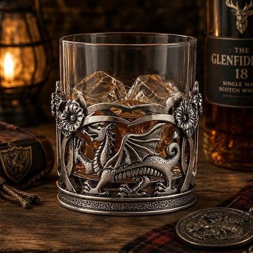 A lowball glass whisky tumbler filled with amber spirits and ice cubes, nestled in an ornate pewter holder featuring Welsh motifs. The front of the holder displays a prominent, detailed relief of the Welsh Dragon (Y Ddraig Goch) with textured scales and open wings, set within an openwork frame topped with sculpted daffodil flowers
