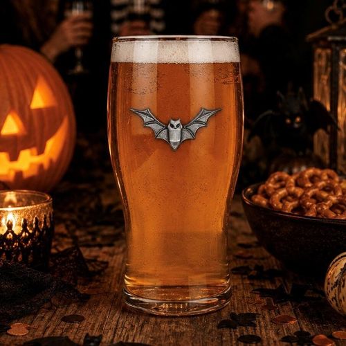 A clear pint beer glass filled with amber ale and a frothy head, featuring a detailed pewter bat motif with outspread wings