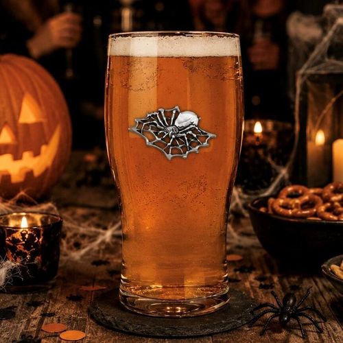 A clear straight pint beer glass filled with golden amber ale and a frothy white head, featuring a detailed pewter spider on a web motif in the center