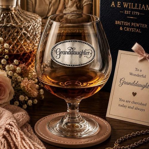 Crystal brandy glass with A E Williams pewter "Granddaughter" emblem, partially filled with amber liquid