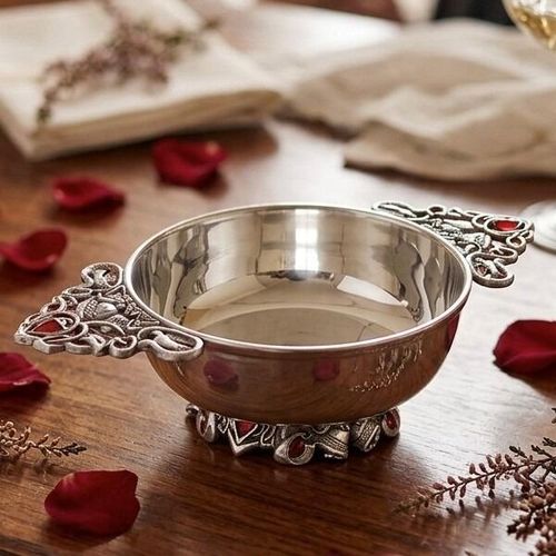 Polished pewter quaich bowl featuring intricate Celtic knotwork handles and a decorative base inlaid with red gemstones, displayed on a wooden table scattered with red rose petals