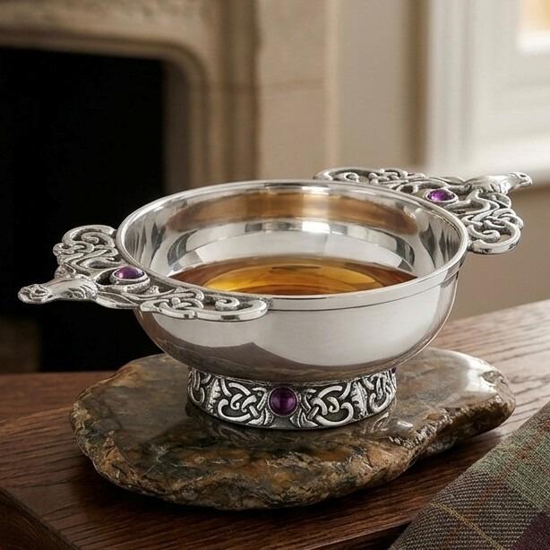Traditional pewter quaich bowl filled with whiskey, featuring ornate Celtic knotwork handles with sculpted stag heads and a decorative base inlaid with purple gemstones, displayed on a flat stone mat