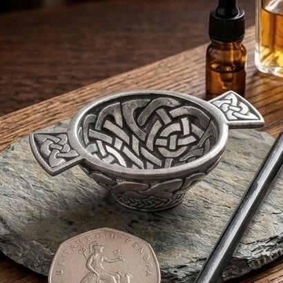 Small antique-finish pewter quaich bowl intricately detailed with raised Celtic knotwork on the interior and handles, displayed on a rough slate mat beside a British fifty pence coin
