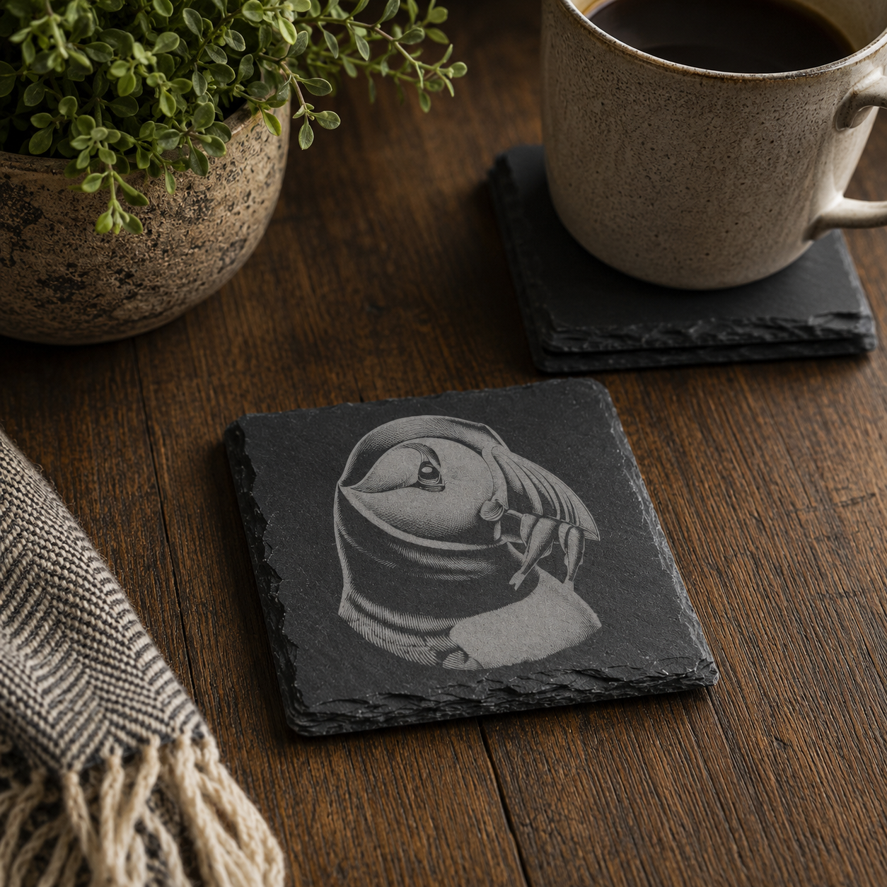 Engraved puffin slate coaster on a rustic wooden table with a coffee mug and houseplant, showcasing a laser-etched puffin design for a Scottish wildlife gift.