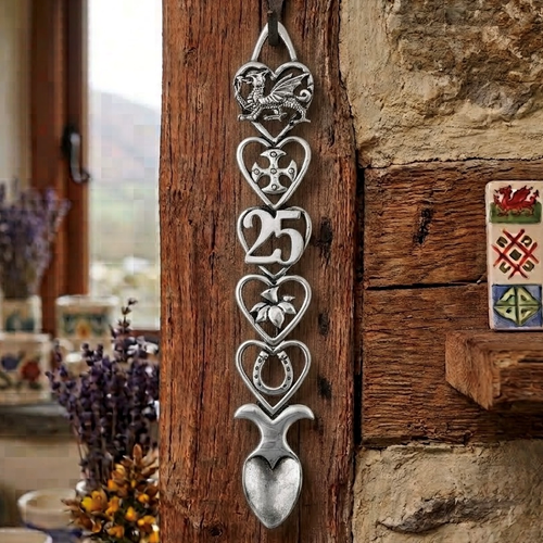 Handcrafted pewter 25th birthday Welsh lovespoon hanging on a rustic wooden beam, featuring detailed symbols including a Welsh dragon, Celtic cross, the number 25, a daffodil, a horseshoe, and a heart-shaped spoon bowl.