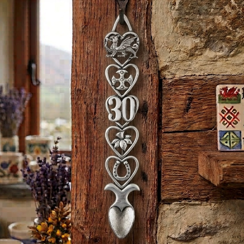 Handcrafted pewter 30th birthday Welsh lovespoon hanging on a rustic wooden beam, featuring detailed symbols including a Welsh dragon, Celtic cross, the number 30, a daffodil, a horseshoe, and a heart-shaped spoon bowl.