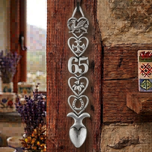 Handcrafted pewter 65th birthday Welsh lovespoon hanging on a rustic wooden beam, featuring detailed symbols including a Welsh dragon, Celtic cross, the number 65, a daffodil, a horseshoe, and a heart-shaped spoon bowl.