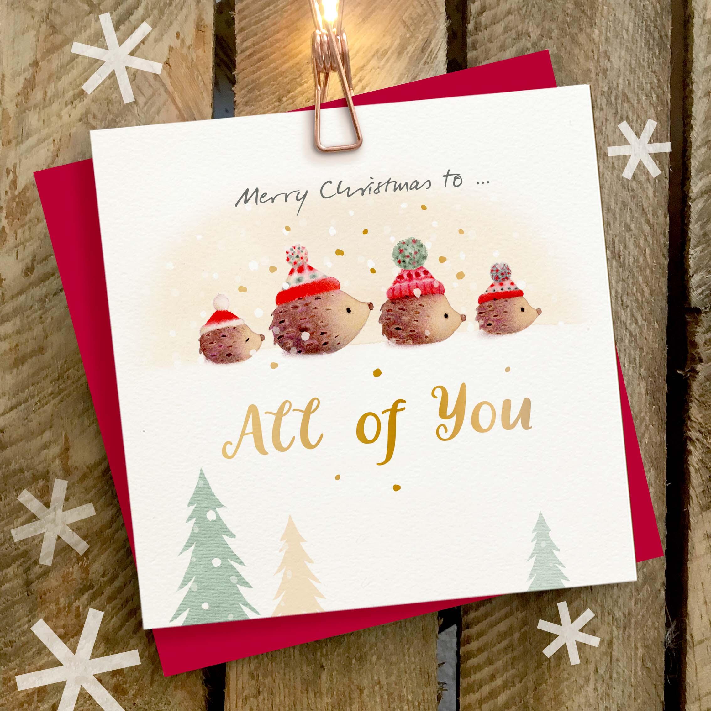 Christmas card featuring a large gold foil ‘All of You’ caption and a family of cute hedgehogs wearing woolly hats.