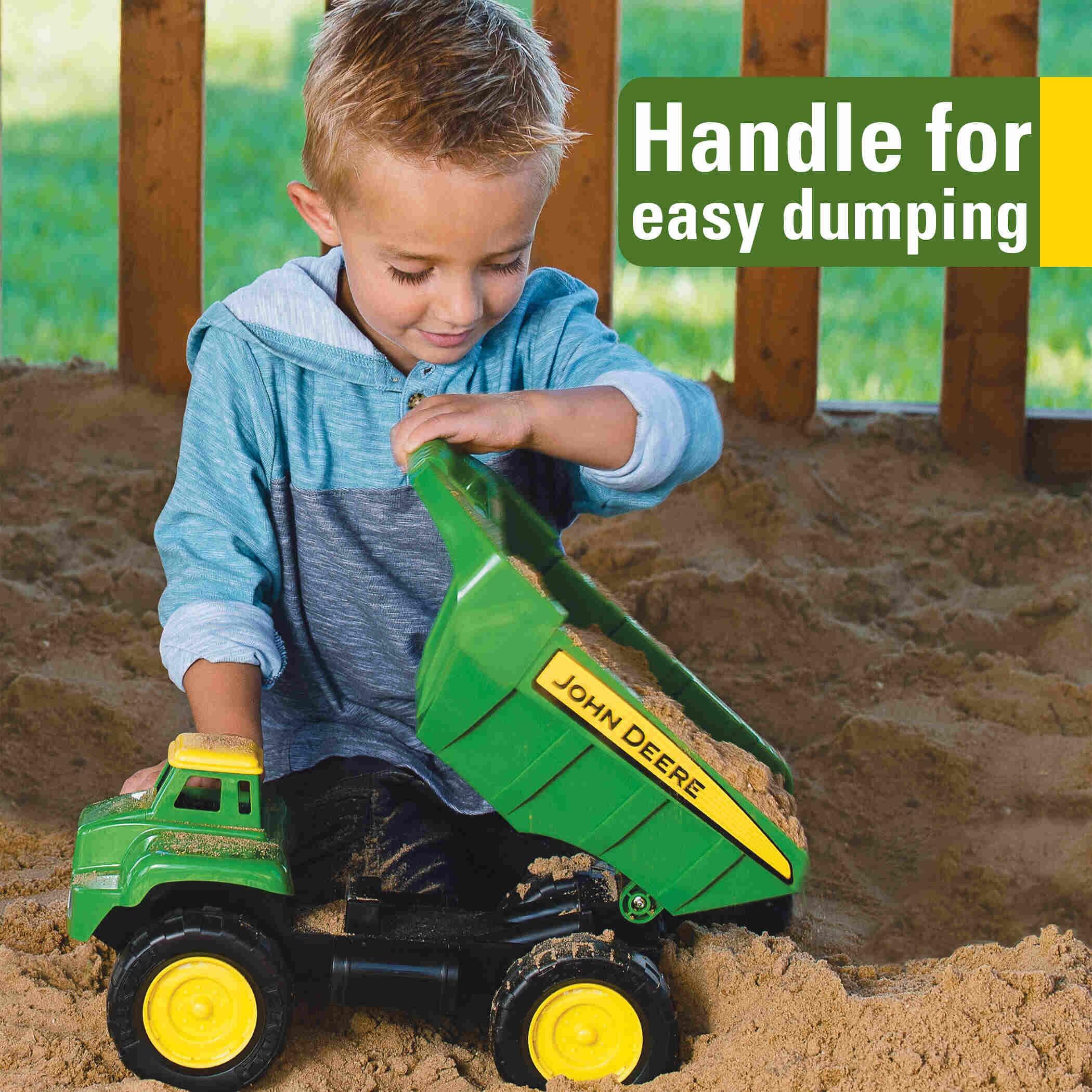 Child playing with a John Deere Big Scoop dump truck toy on sand, using the dumping handle.