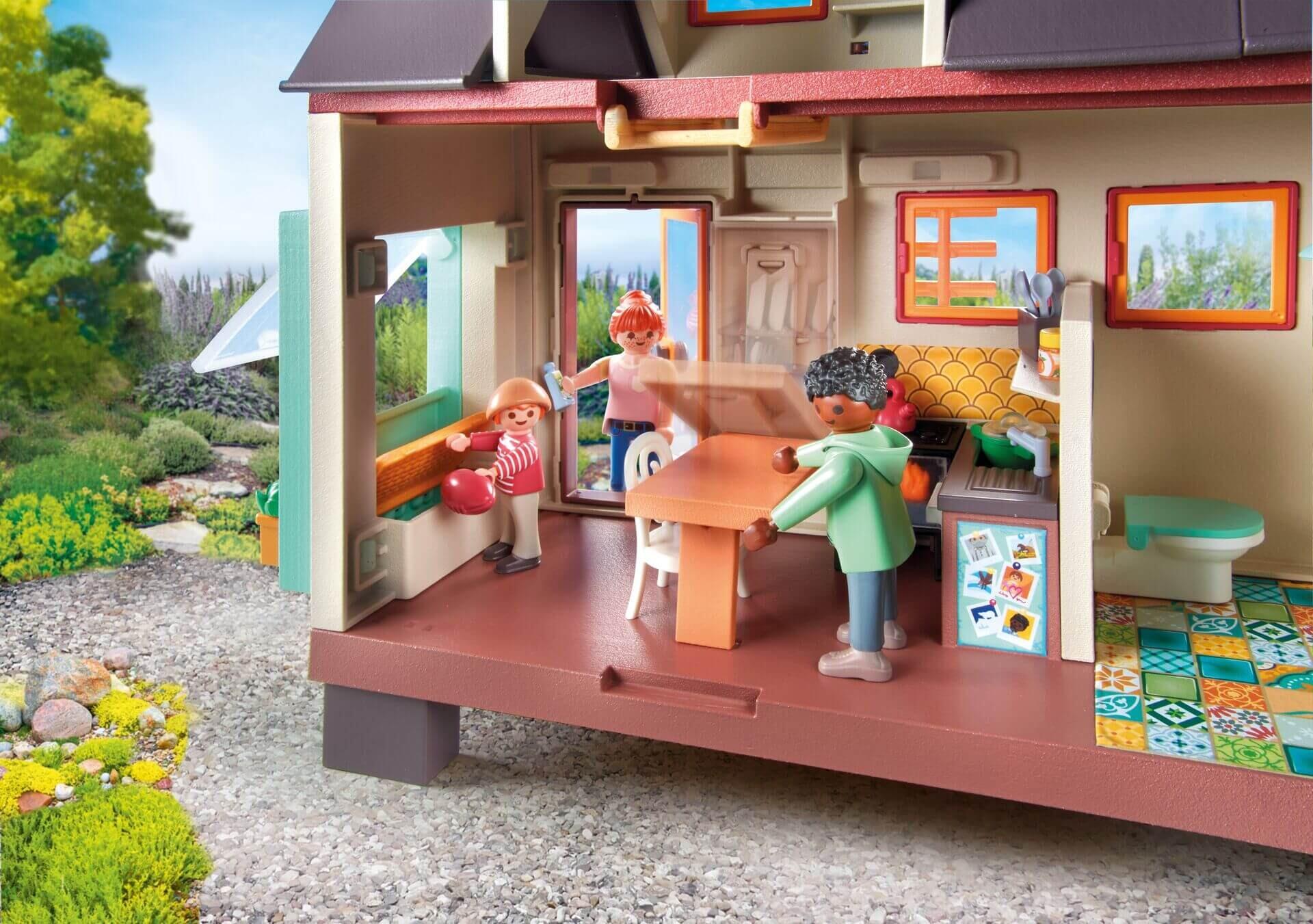 Interior of the PLAYMOBIL Tiny House showing three figures inside the kitchen and dining area, with one adjusting the folding table and another child standing by the open wall bench.