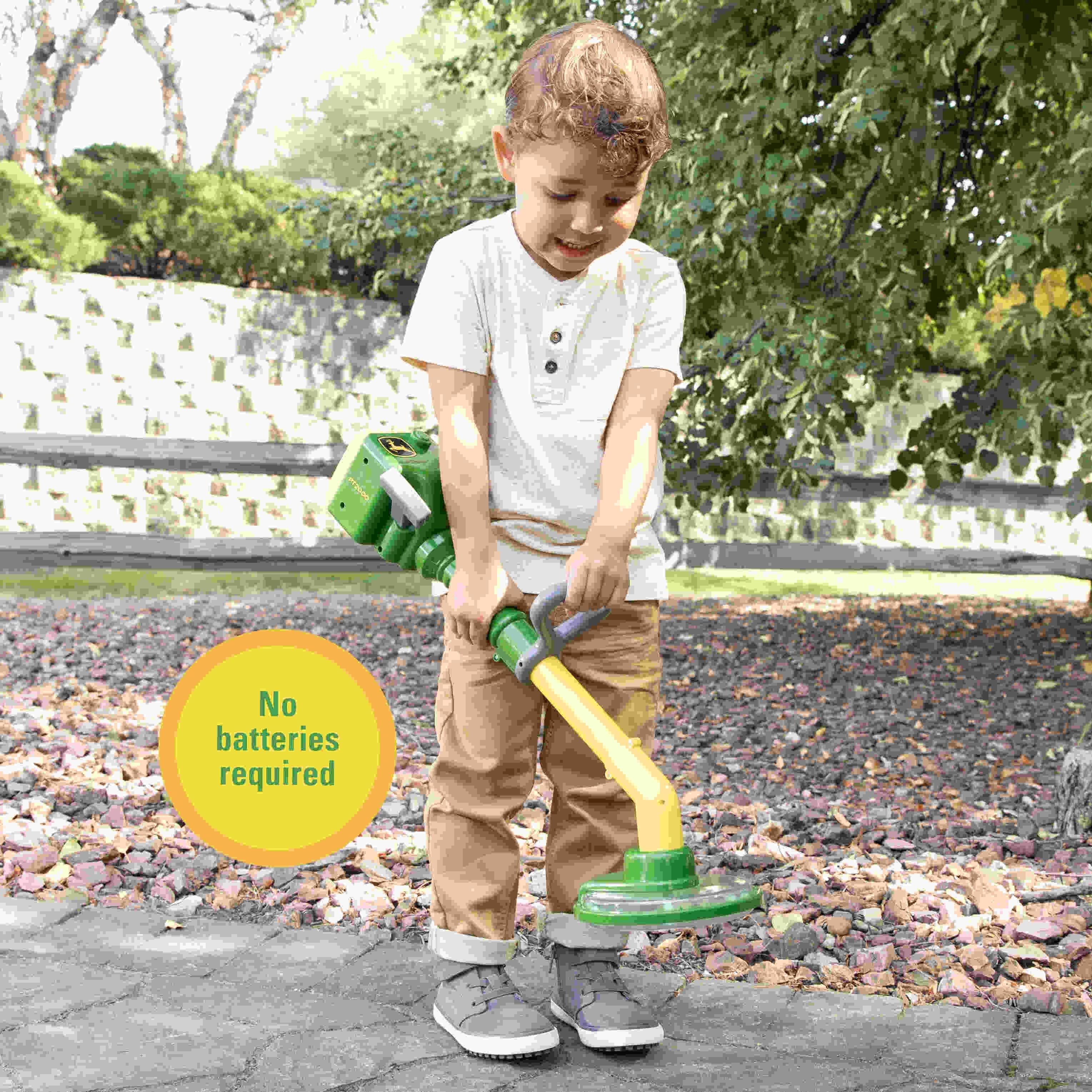 hild holding and using a John Deere toy power trimmer outdoors on a pebble path.