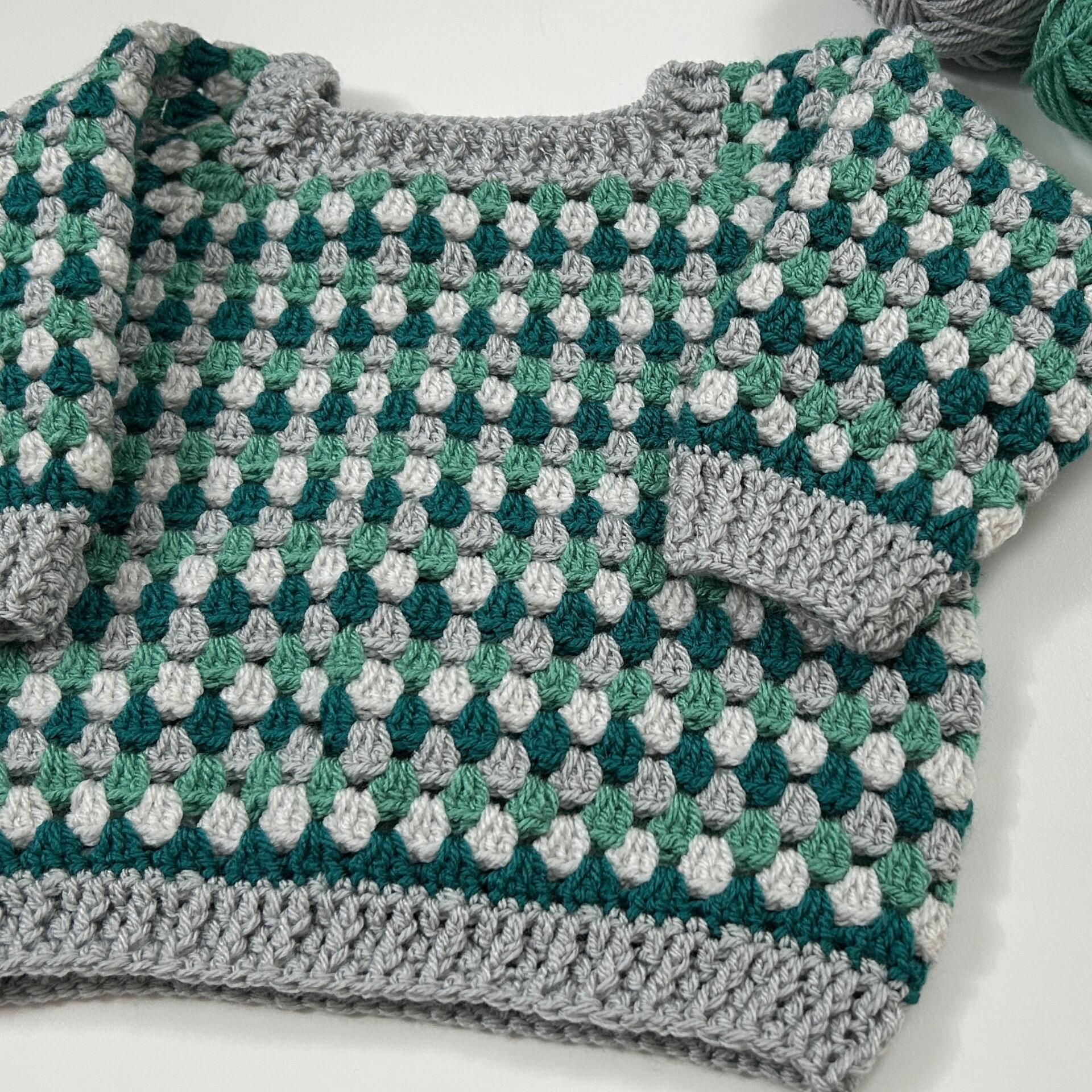 A cosy crocheted toddler sweater featuring a classic granny stitch pattern in shades of grey, green, and white, with ribbed cuffs and edging.