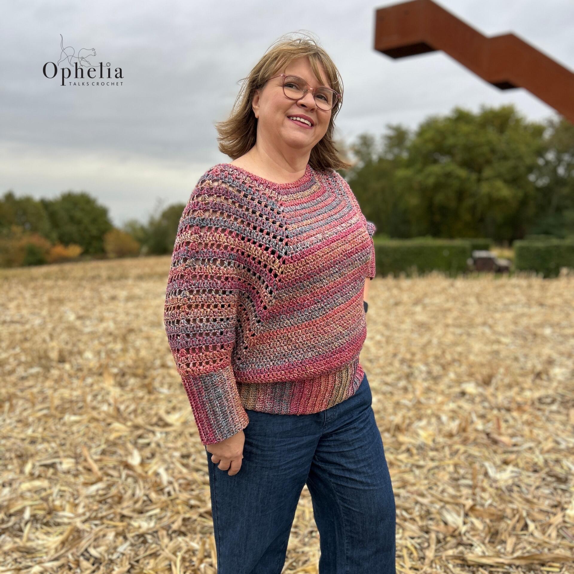 Woman wearing a crochet batwing sweater in pink and mauve shades, Impressions Renoir, by Ophelia Talks Crochet, smiling outdoors in a field setting.