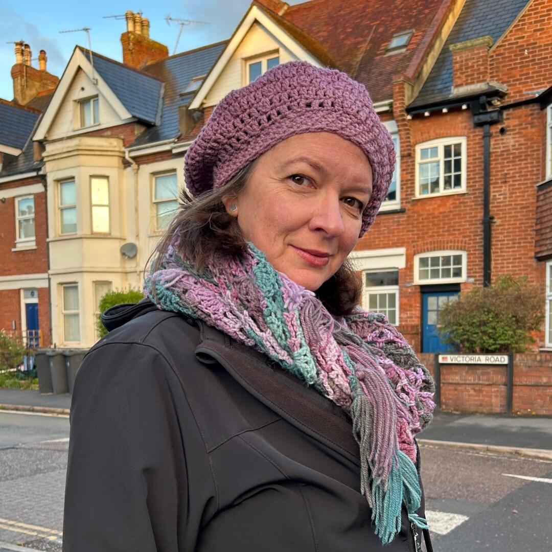 Woman wearing a purple crochet beret and multicoloured crochet scarf, standing on Victoria Road with traditional British houses in the background.