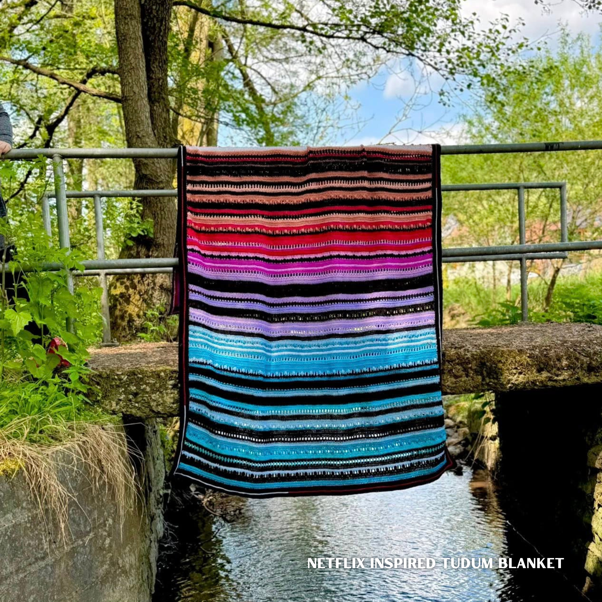 Netflix-inspired Tudum crochet blanket displayed over a metal railing above a stream, featuring bold horizontal stripes in black, red, pink, purple, turquoise and blue, photographed outdoors in a leafy woodland setting