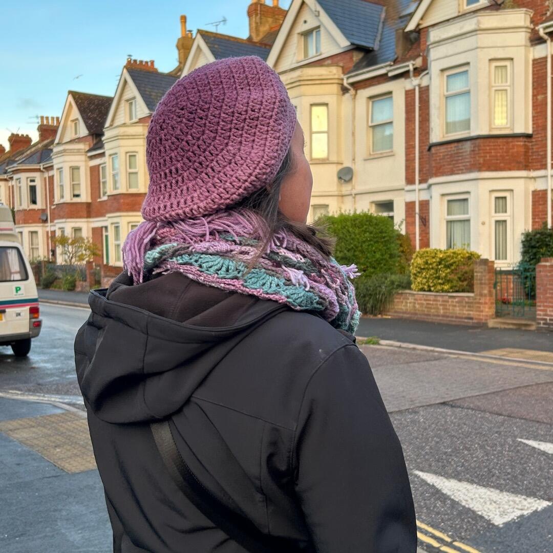 Back view of a woman wearing a purple crochet beret and textured multicoloured crochet scarf, walking along a residential street lined with Victorian-style houses.