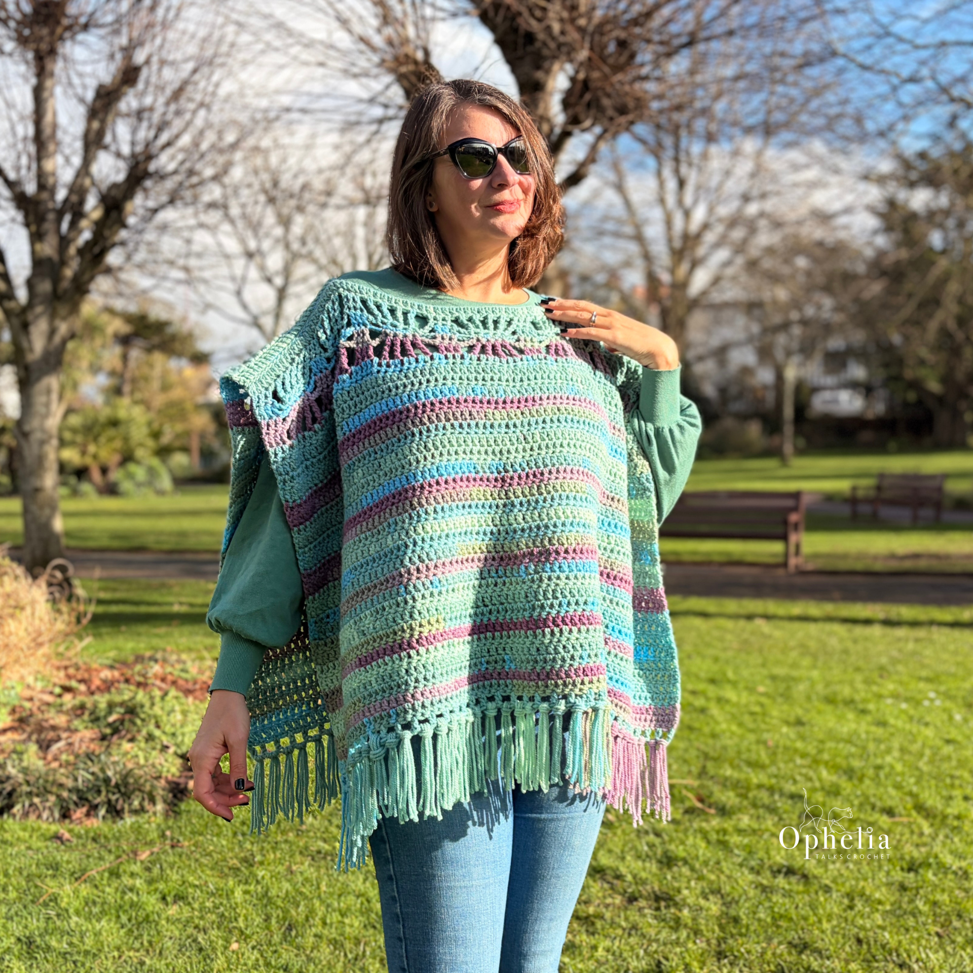 Woman wearing the Garden Slate Poncho crochet design by Ophelia Talks Crochet in a sunny park. The ruana style chunky crochet poncho features soft mint, teal and purple tones with tassel fringe, styled over jeans and a green sweater.