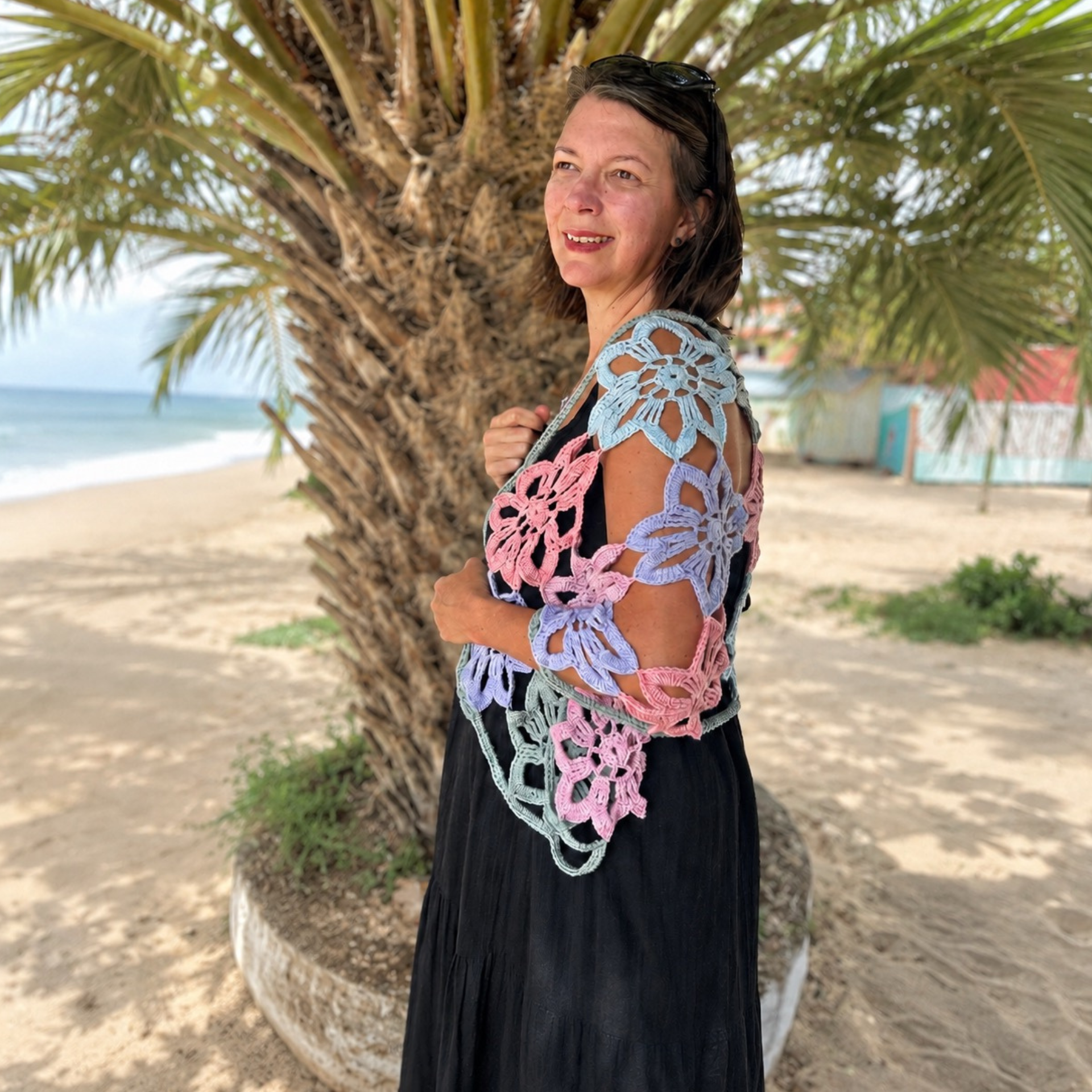Model wearing a pastel crochet flower shawl over a black dress, standing on a sandy beach beside a palm tree with the sea in the background.