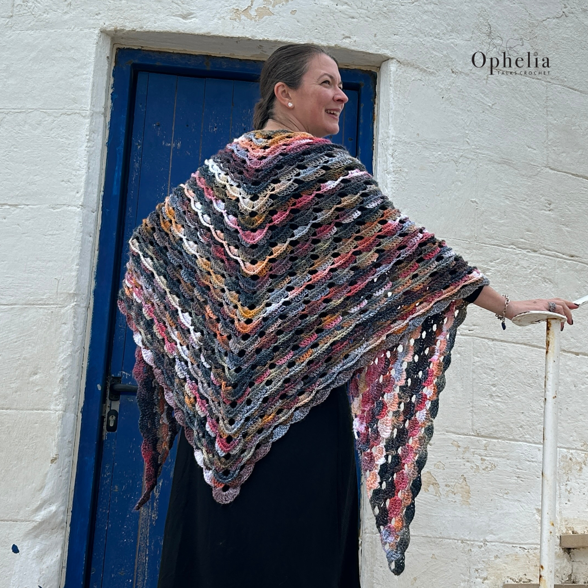 Woman modelling the multicoloured crochet Paphos Shawl in shades of pink, grey, cream and tan, standing beside a blue door against a whitewashed wall.