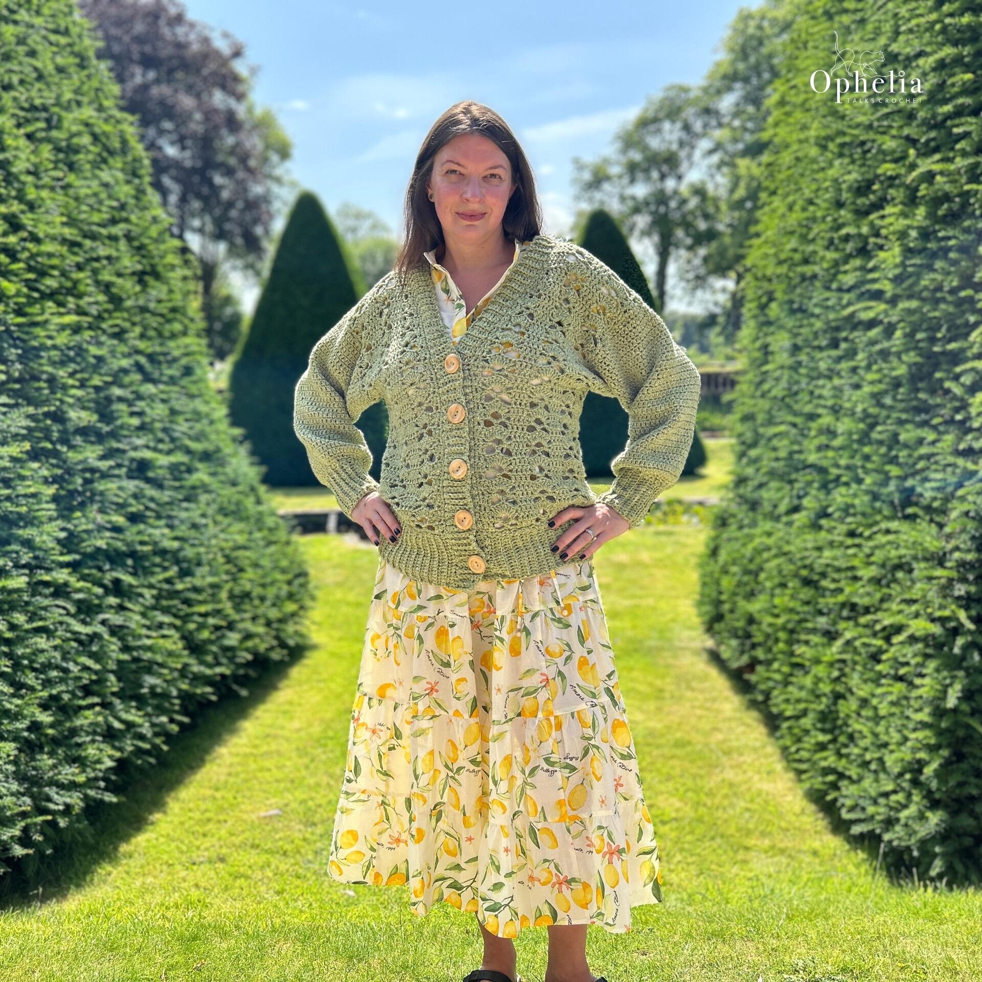 A woman stands confidently in a sunlit formal garden, wearing a hand-crocheted sage green cardigan with large wooden buttons over a white dress patterned with yellow lemons and green leaves. Tall, manicured evergreen trees surround her, and the sky is clear and blue. The Ophelia Talks logo is visible in the top right corner.