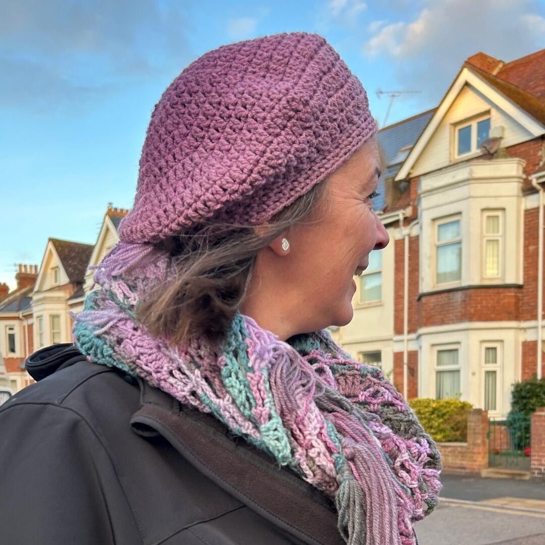 Side view of a woman wearing a handmade purple crochet beret and a pastel multicoloured crochet scarf, standing in front of traditional British homes.