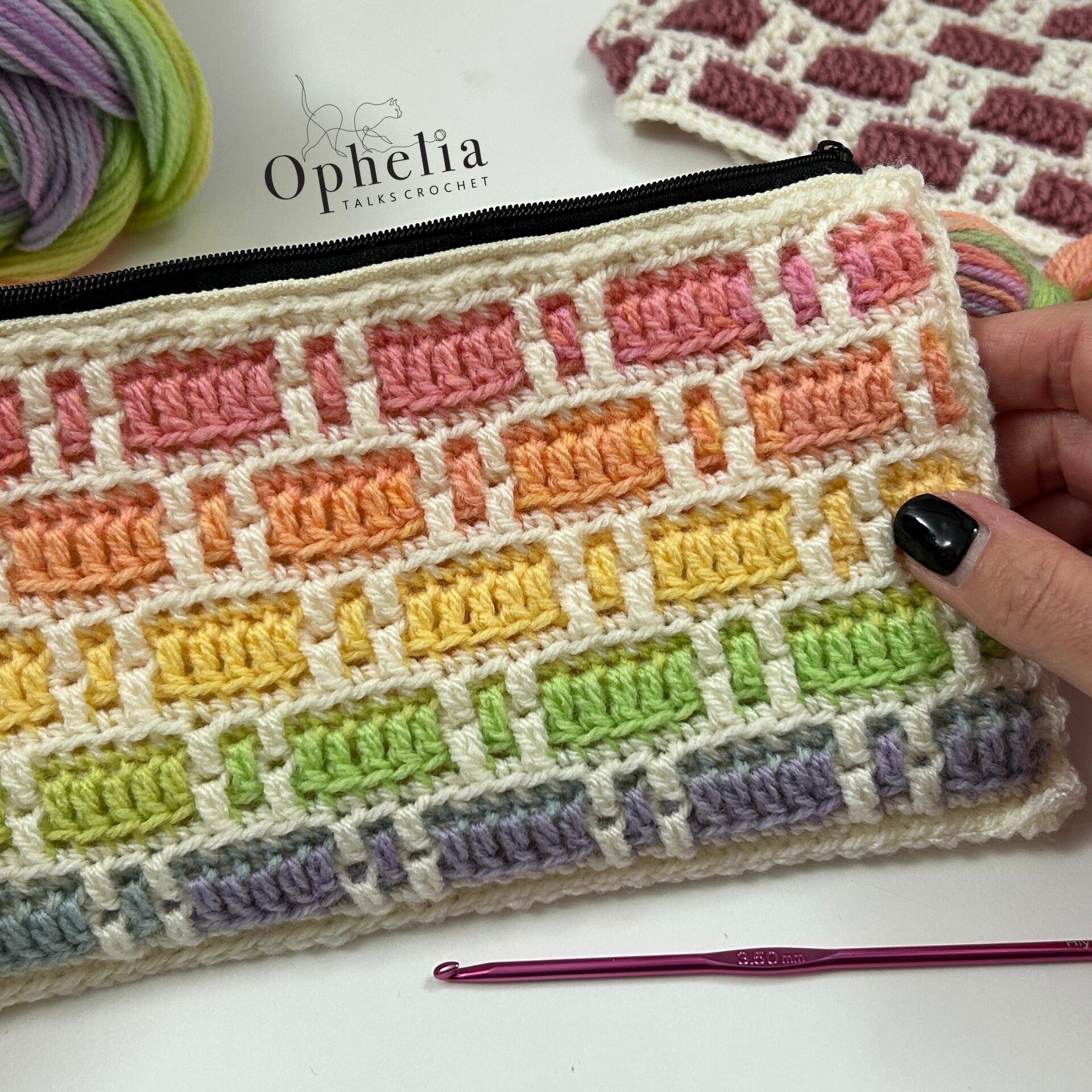 A close-up of a crocheted zipper pouch featuring a colorful brick-like pattern with pastel shades of pink, orange, yellow, green, and purple, separated by cream-colored stitches. The pouch is held by a hand with black nail polish, and a crochet hook lies beside it. The background shows yarn balls and the Ophelia Talks Crochet logo.