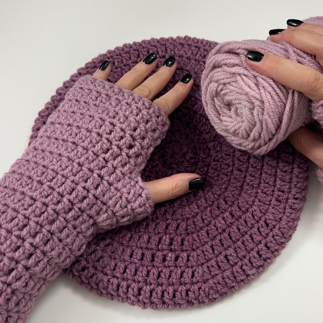 Close-up of handmade purple crochet fingerless gloves and matching beret laid on a table, with hands wearing the gloves and holding a ball of yarn.
