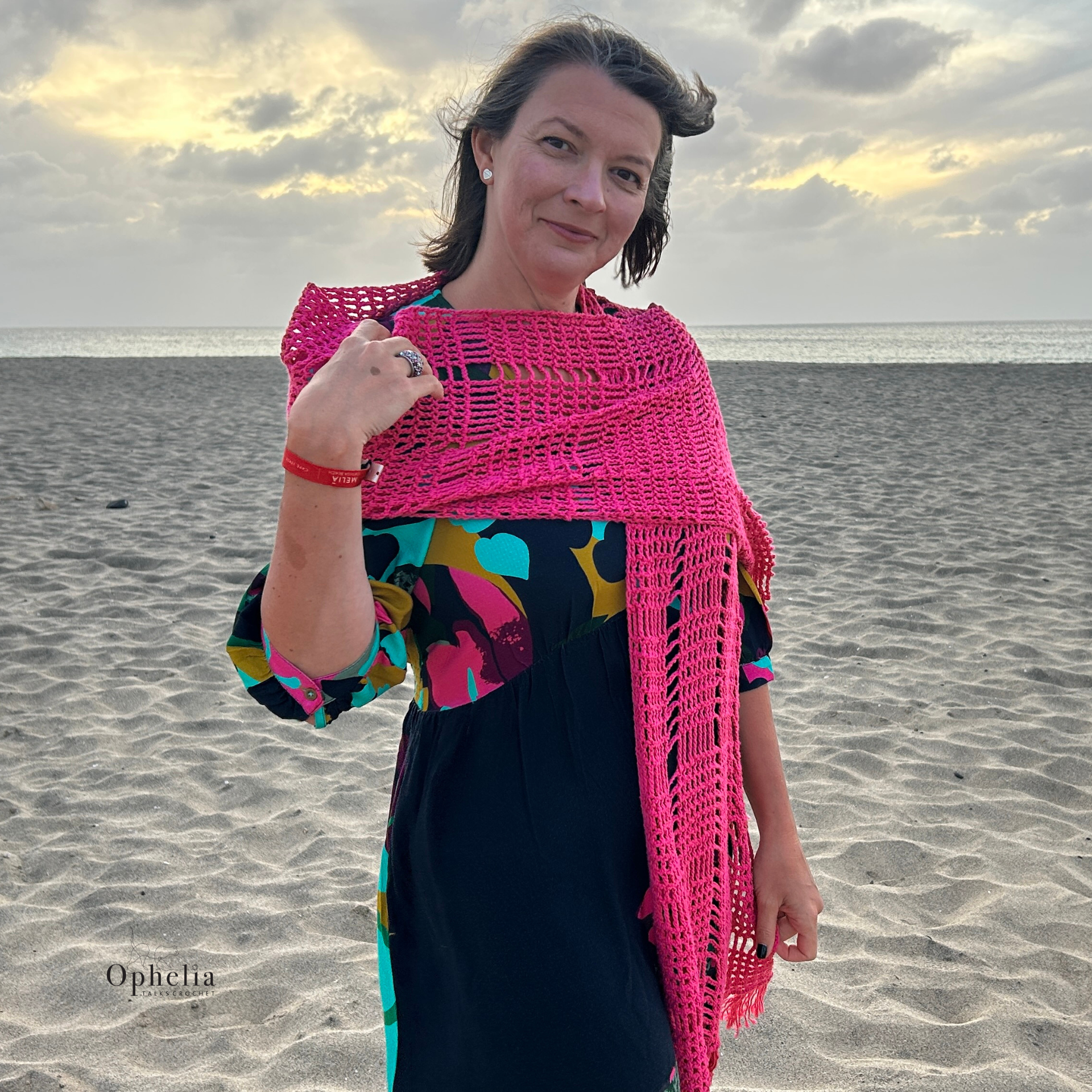 Woman wearing bright raspberry pink crochet shawl with geometric box pattern draped across shoulders on sandy beach at sunset, Ophelia Talks Crochet design