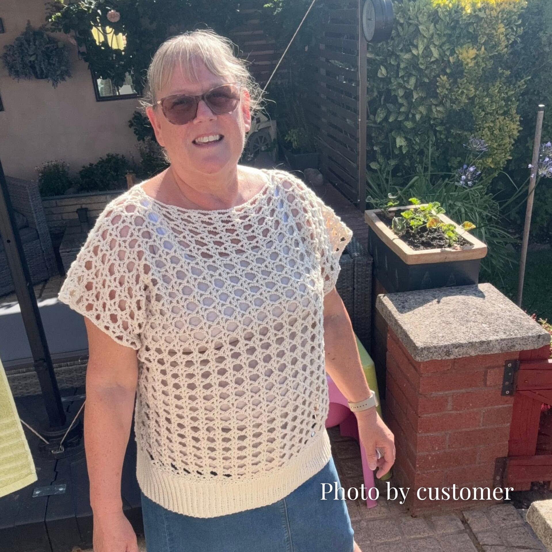 Front view of a smiling woman wearing sunglasses modeling a cream-coloured Daydream summer crochet top—made in Wendy Supreme Cotton Love yarn in the shade Linen—with airy openwork stitches, ribbed hem and short sleeves, standing in a sunny garden patio.