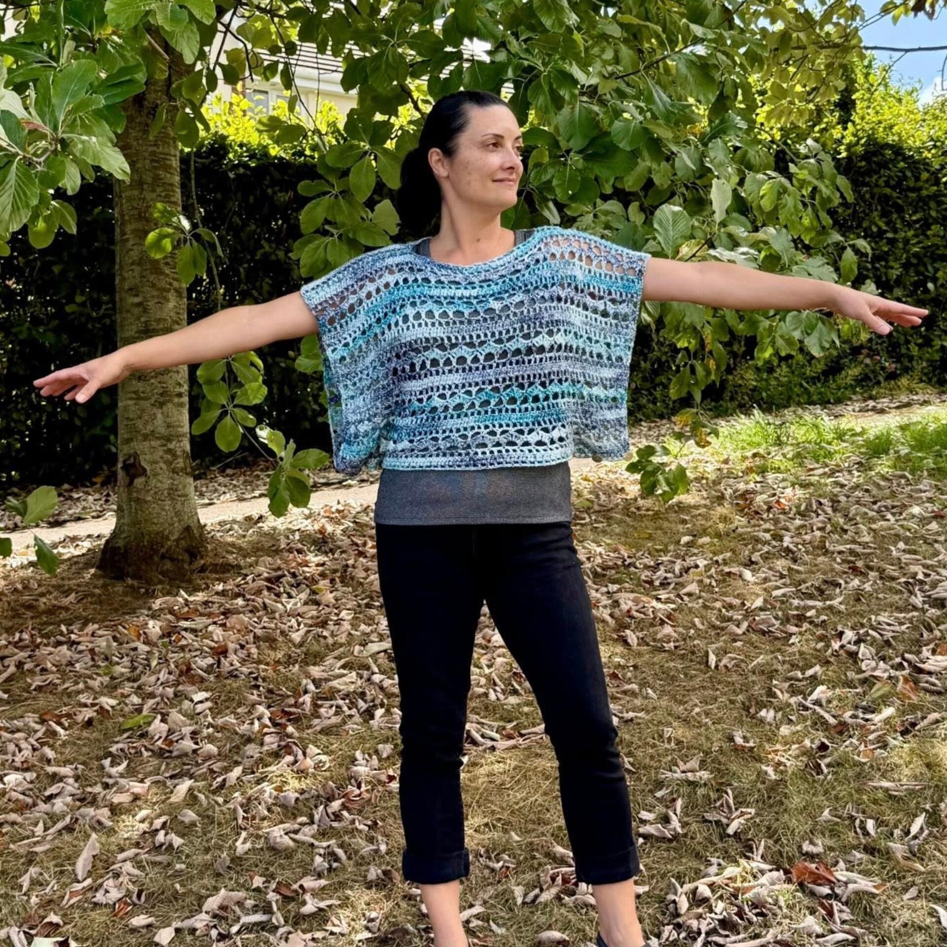 Woman standing outdoors with arms outstretched, wearing the Pixie Bolero — a loose-fitting, lacy crochet top in shades of blue and turquoise, layered over a grey tank top. She is barefoot on dry grass with scattered leaves, with a large green-leaved tree in the background.