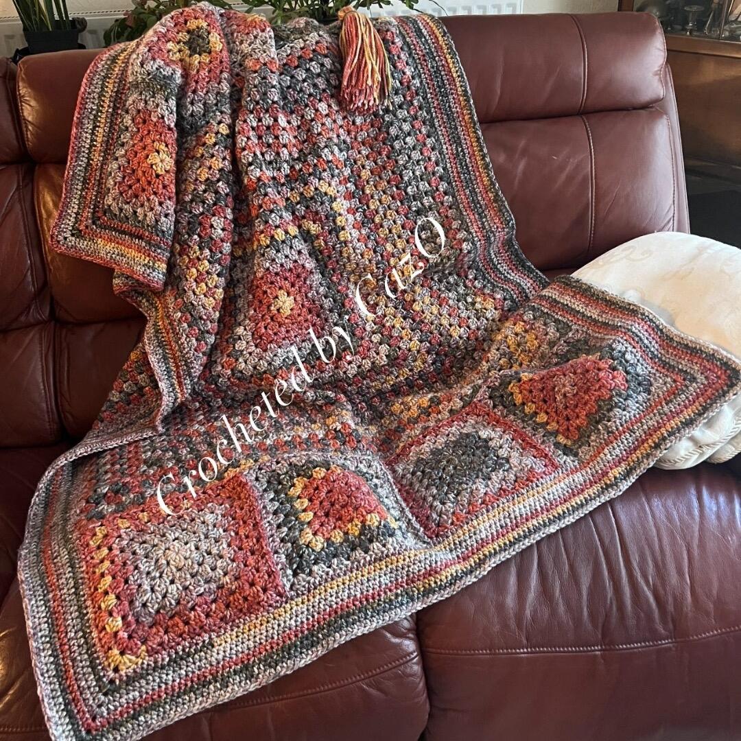 A crocheted “Crocus Blanket” in the Hedgerow range’s Conker colourway draped over a brown leather sofa. The blanket features rich autumnal tones of rust, gold, grey, and warm browns, arranged in traditional granny squares with a textured, multi-striped border. A matching tassel rests on top of the blanket, showcasing the cosy, handmade finish.