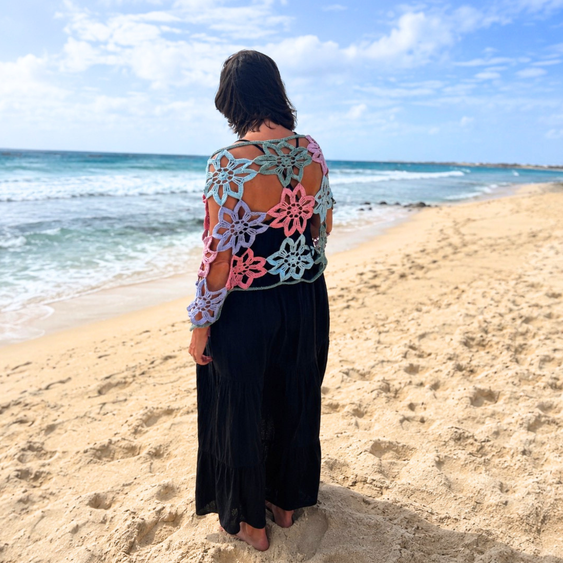 Model wearing a pastel crochet flower shawl over a black dress while walking along a sandy beach with ocean waves and blue sky in the background.