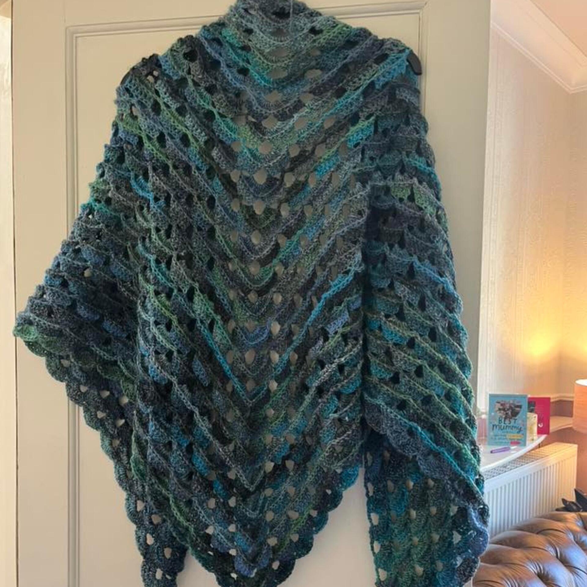 Crochet Paphos Shawl made in James C. Brett Emotions DK shade Calm (EM07), featuring a large triangular shape with layered openwork lace and scalloped edging in blended shades of teal, blue, green and soft grey, draped over a door indoors.