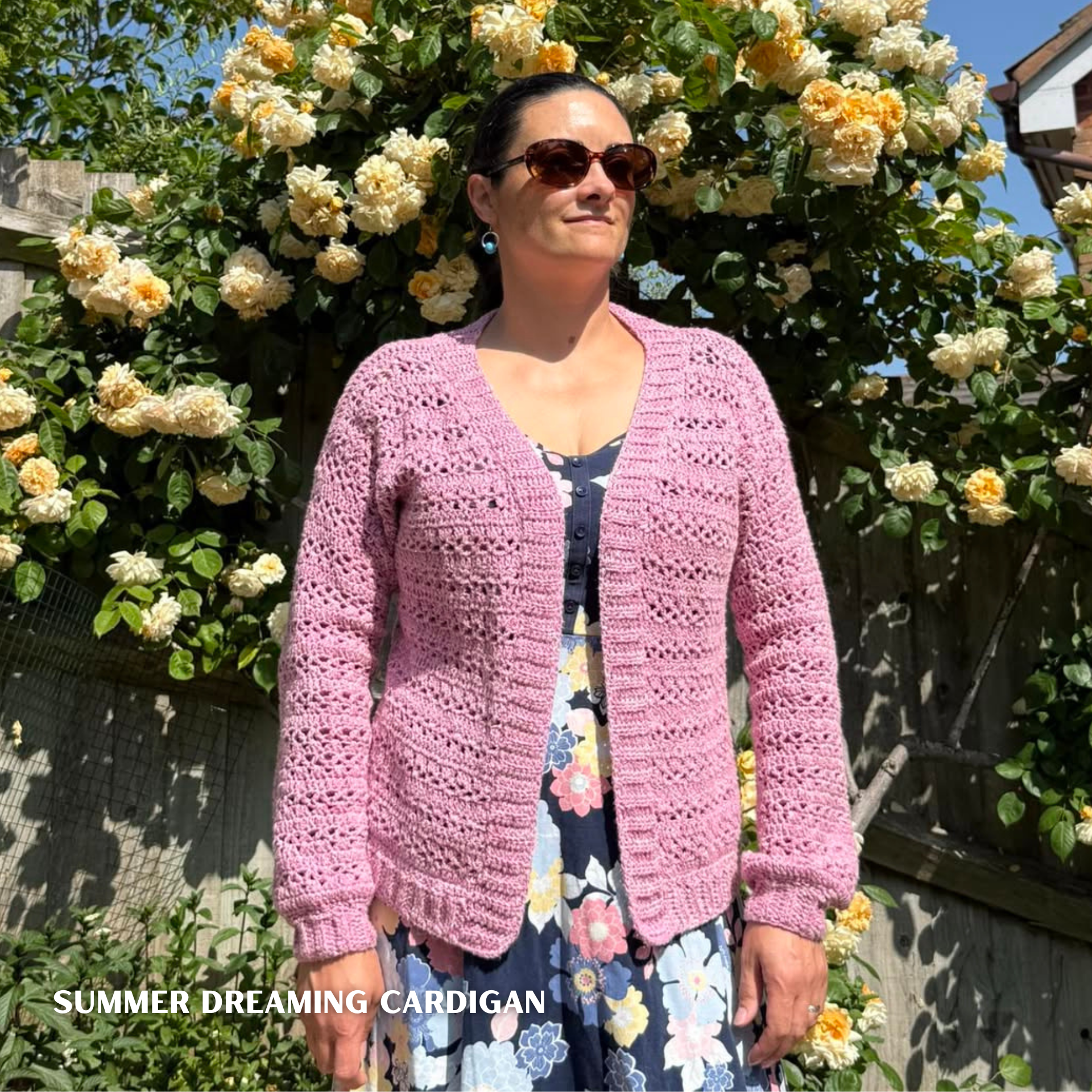 crochet cardigan in King Cole Pink Denim worn by a lady in a blue dress in front of a rose garden
