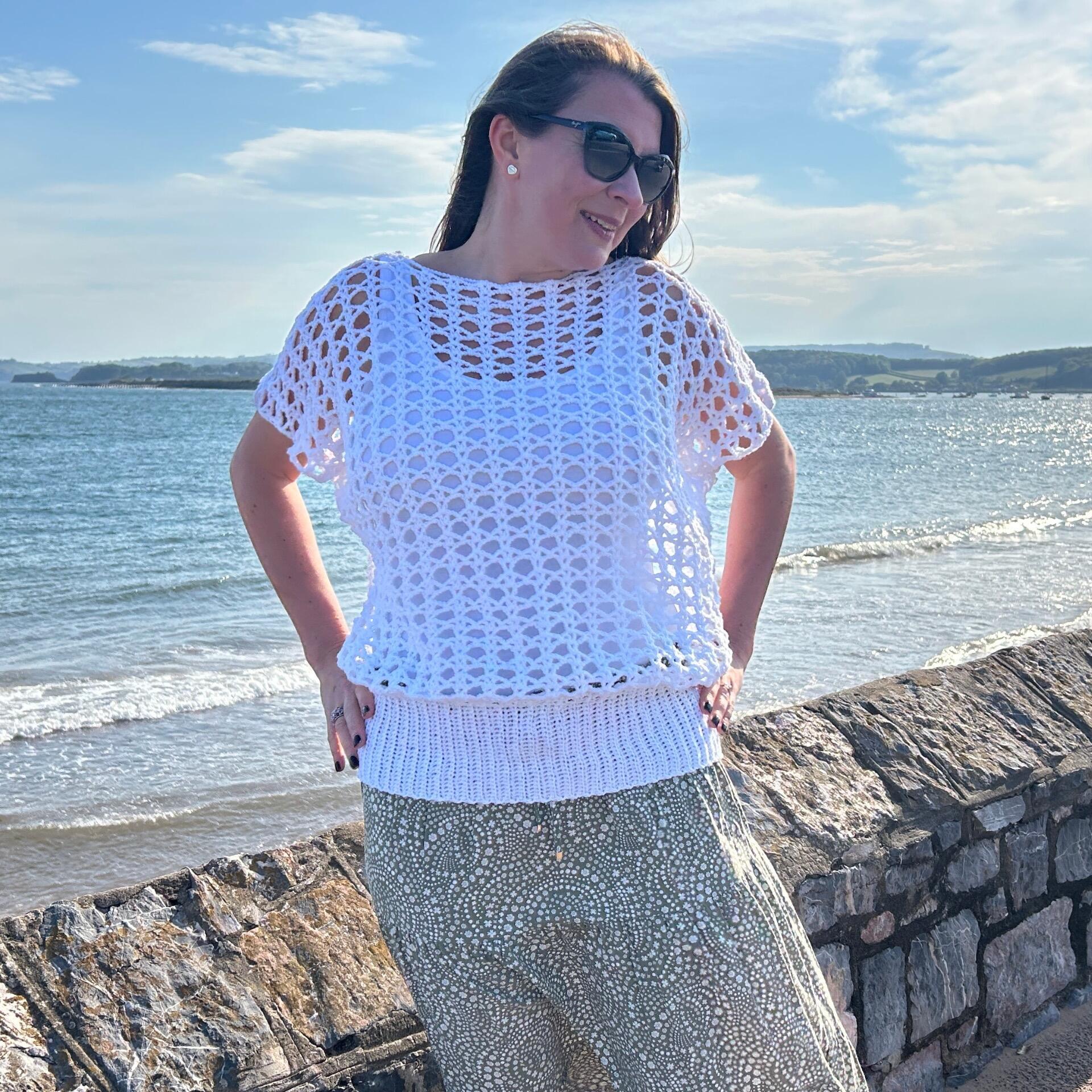A woman wearing sunglasses poses on a stone walkway by the sea, smiling in a white crocheted top with a ribbed waistband and short sleeves. The sky is clear with soft clouds, and the water gently laps against the shore.