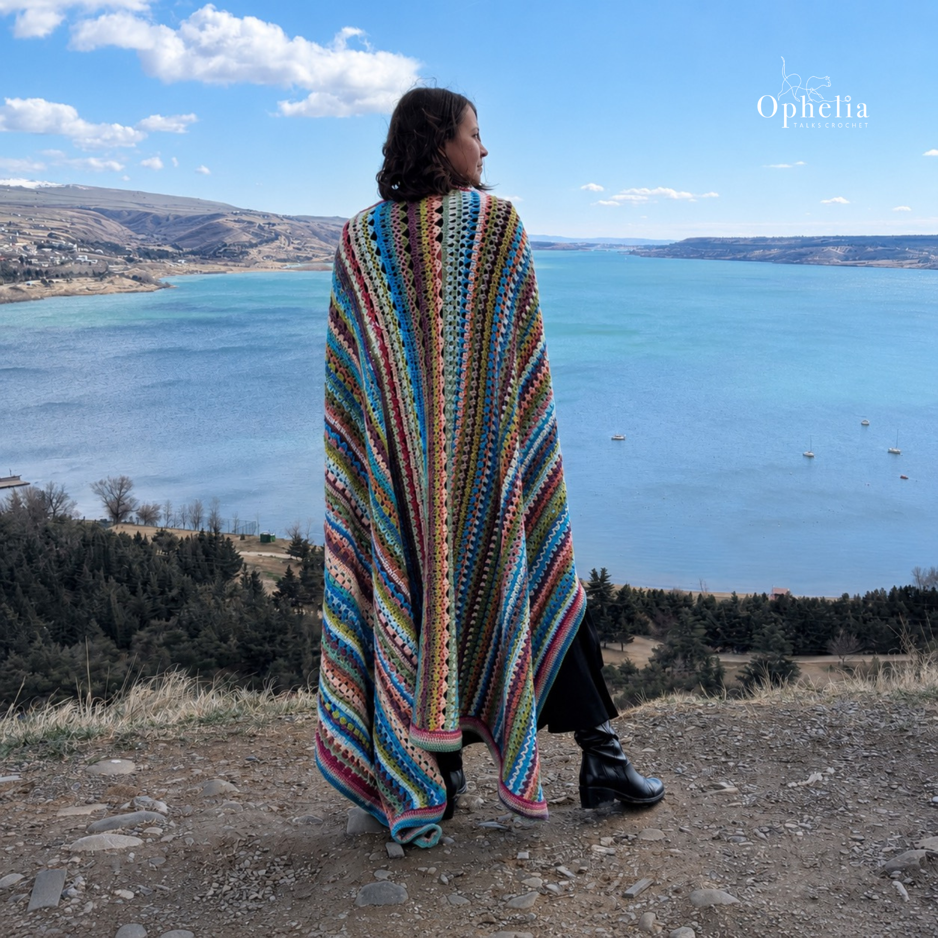 Woman wearing a colourful striped crochet blanket wrap, standing on a hilltop viewpoint overlooking a bright blue lake, distant hills and a wide sky with soft clouds.