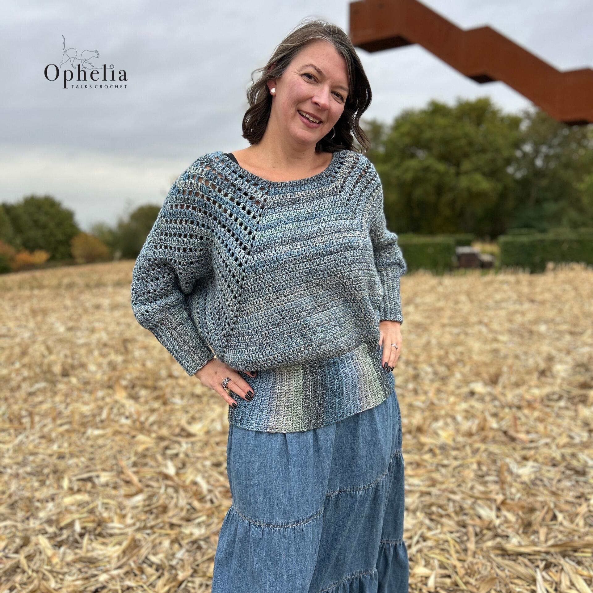 Woman wearing a handmade crochet batwing sweater in blue tones by Ophelia Talks Crochet, standing in a field near the Vlooybergtoren in Belgium