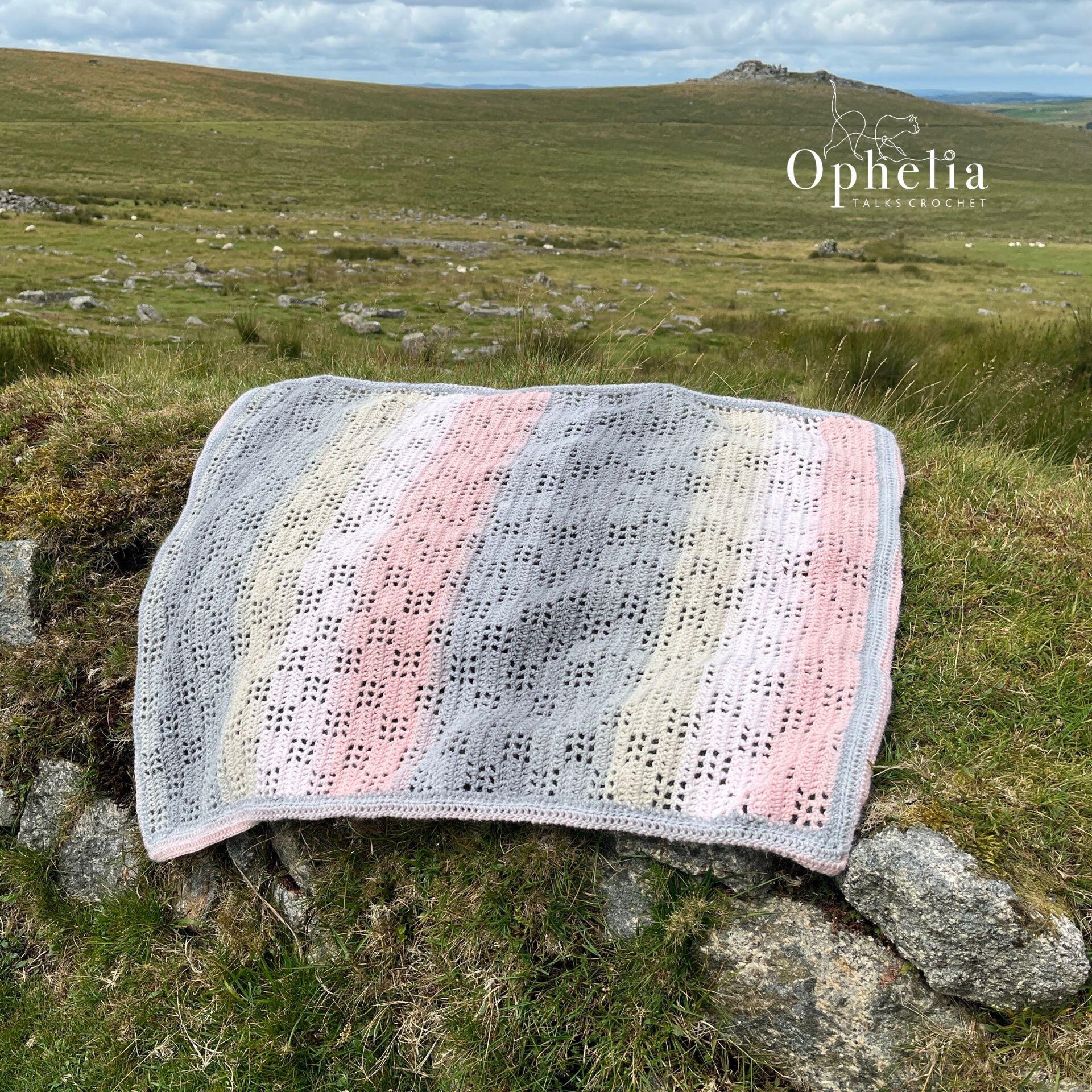 Filet crochet baby blanket in pastel shades of pink, grey, and cream displayed on grassy moorland rocks. Handmade using the Ode To Baby crochet pattern by Ophelia Talks Crochet