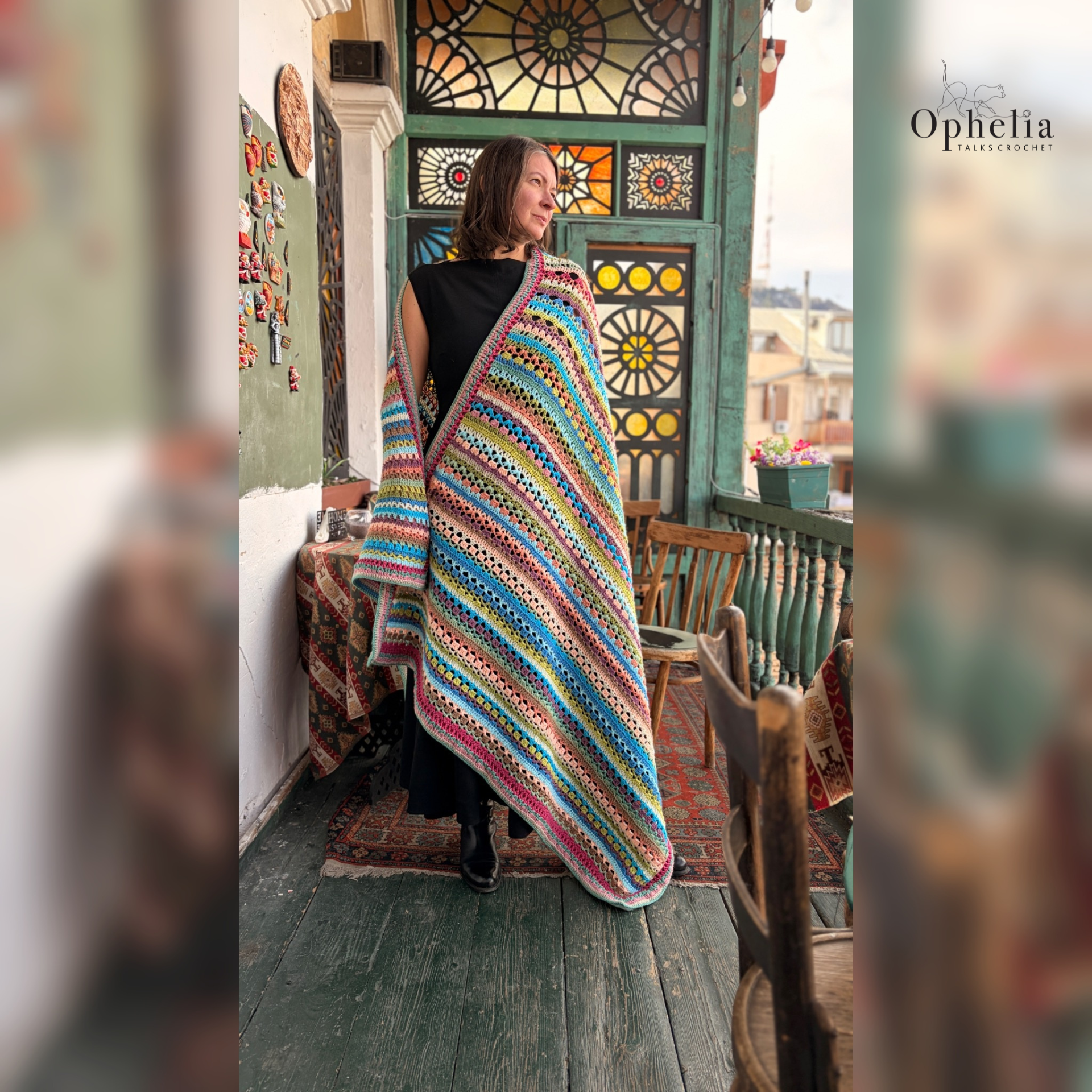 Woman modelling a colourful striped crochet blanket wrap on a vintage balcony with stained-glass windows, patterned rugs, wooden chairs and traditional Georgian-style décor