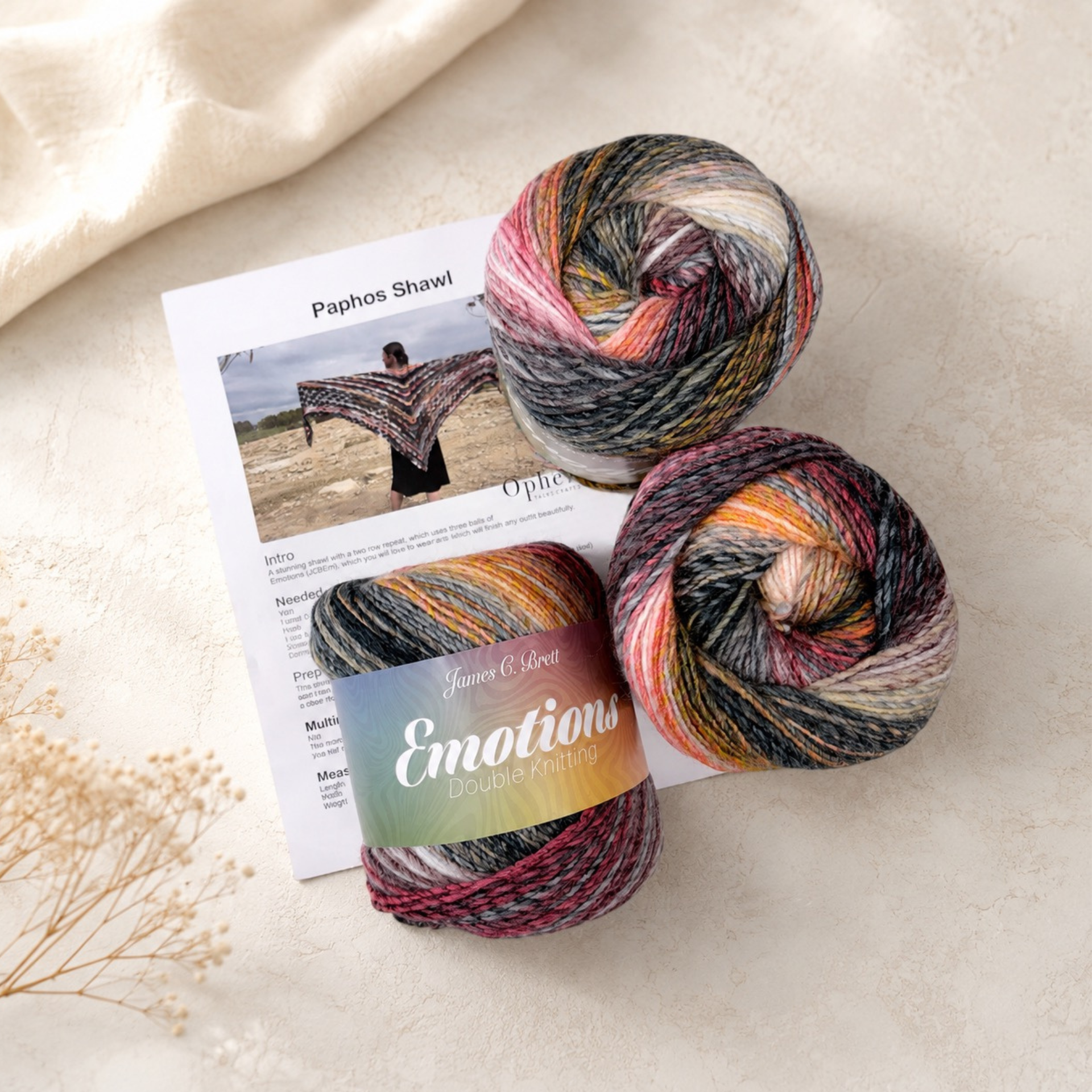 Paphos Shawl yarn pack featuring three balls of James C. Brett Emotions DK yarn in shade EM10 Interest with the printed crochet pattern, arranged on a soft cream textured background.