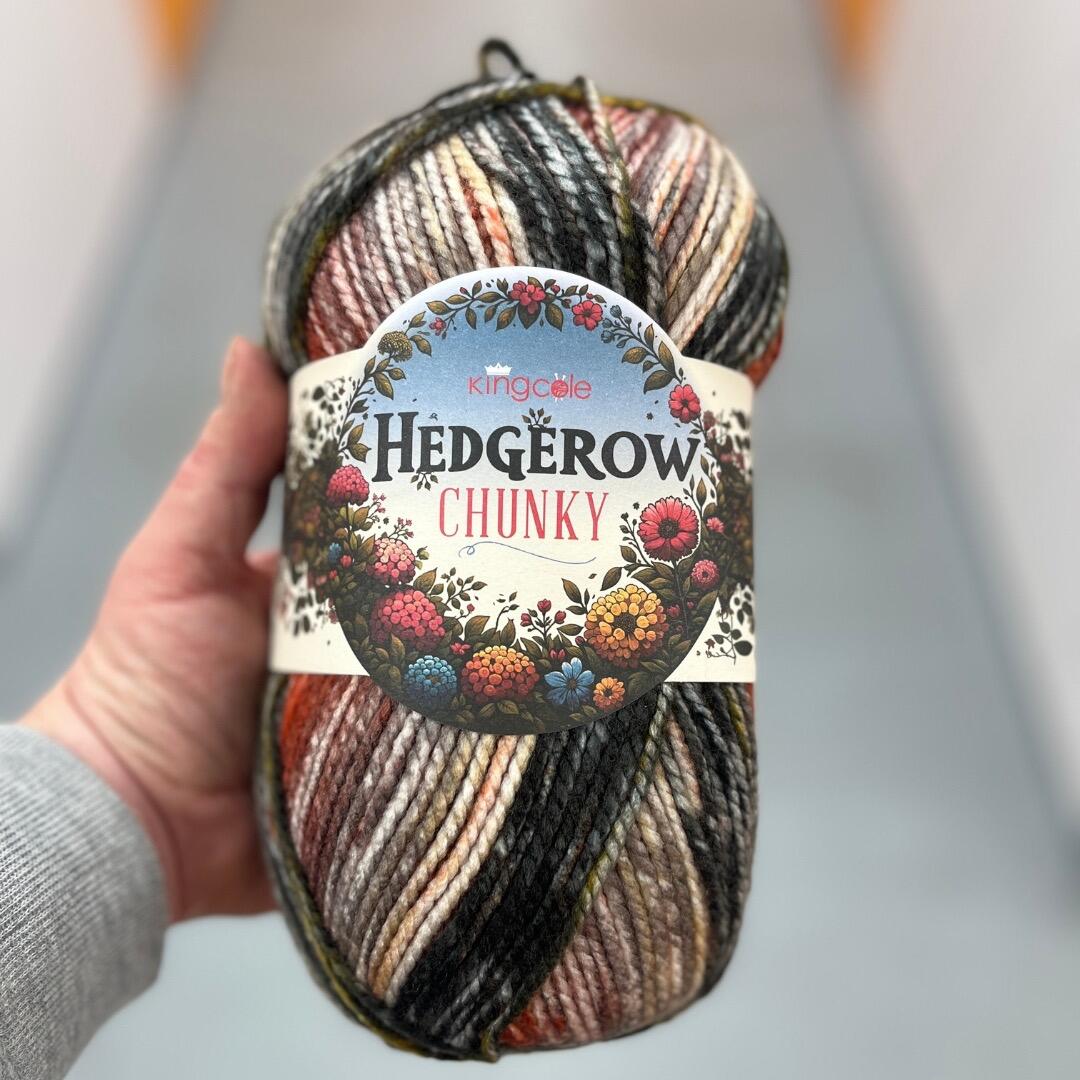 A hand holding a ball of King Cole Hedgerow Chunky yarn in the colour Conker. The yarn features warm autumnal variegated shades of brown, orange, cream, and black. The label shows the Hedgerow Chunky branding with a floral design