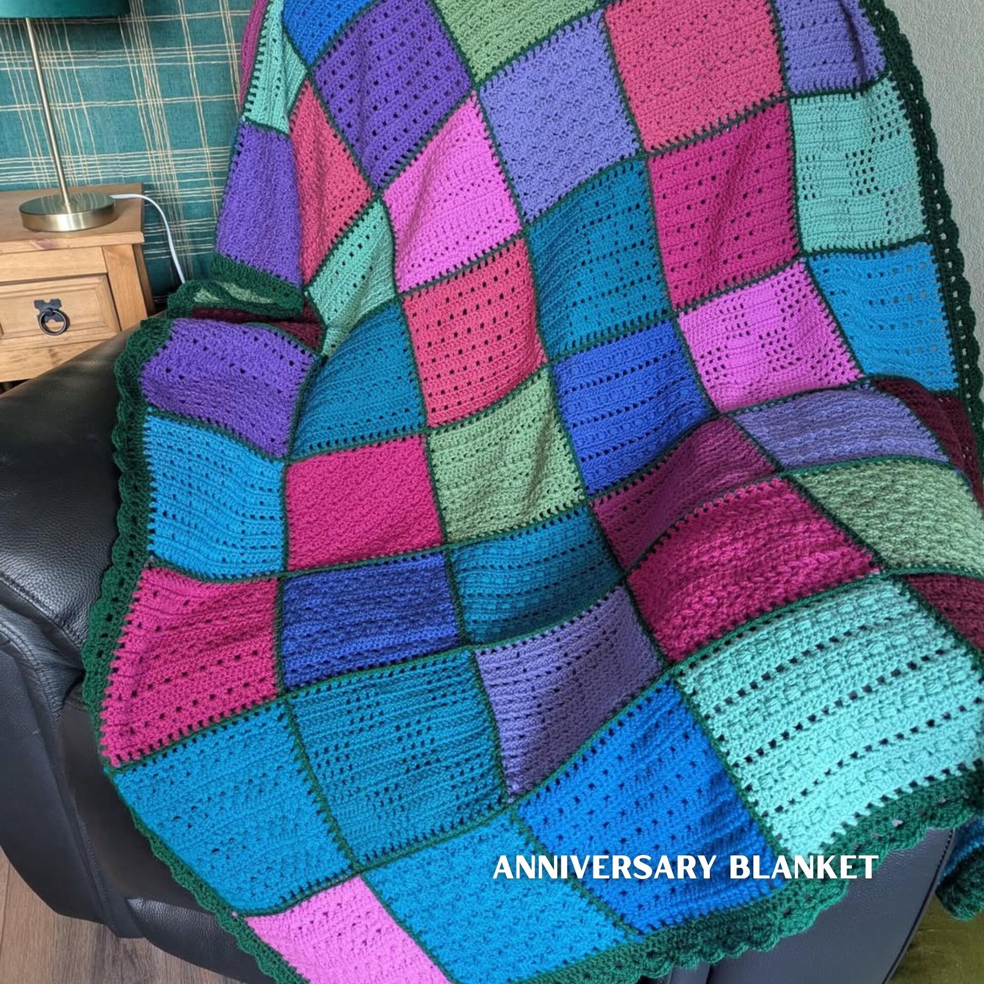 Crocheted Anniversary Blanket made with the Enchanted Forest yarn pack, draped over an armchair, featuring textured crochet squares in rich teal, turquoise, pink, purple and forest green tones, finished with a dark green scalloped border for a vibrant patchwork effect.