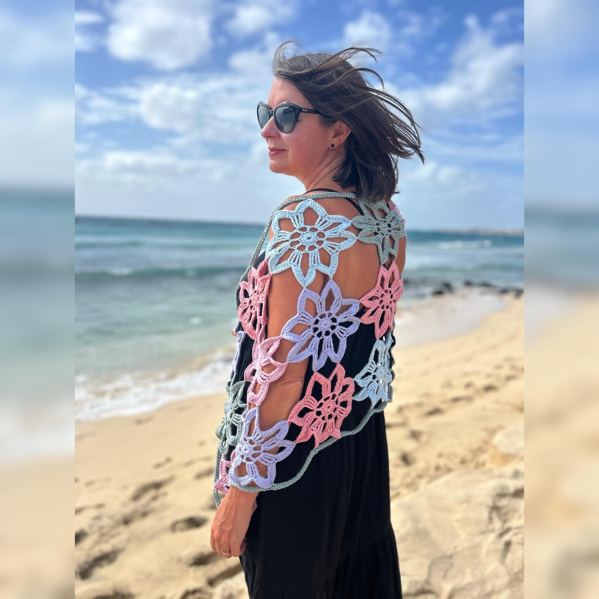 Model wearing the Flower Power crochet shawl on a sandy beach, with pastel flower motifs in blue, pink, lilac and sage draped over a black dress against a soft ocean and sky background.